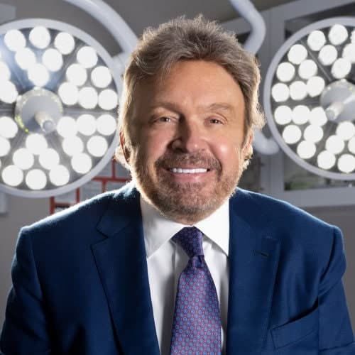 Nip Tuck Aesthetics surgeon in an operating room with surgical lighting Professional plastic surgeon at Nip Tuck Aesthetics clinic offering cosmetic procedures Accurate image description showing a confident surgeon in a healthcare setting Relevance: Relevant.