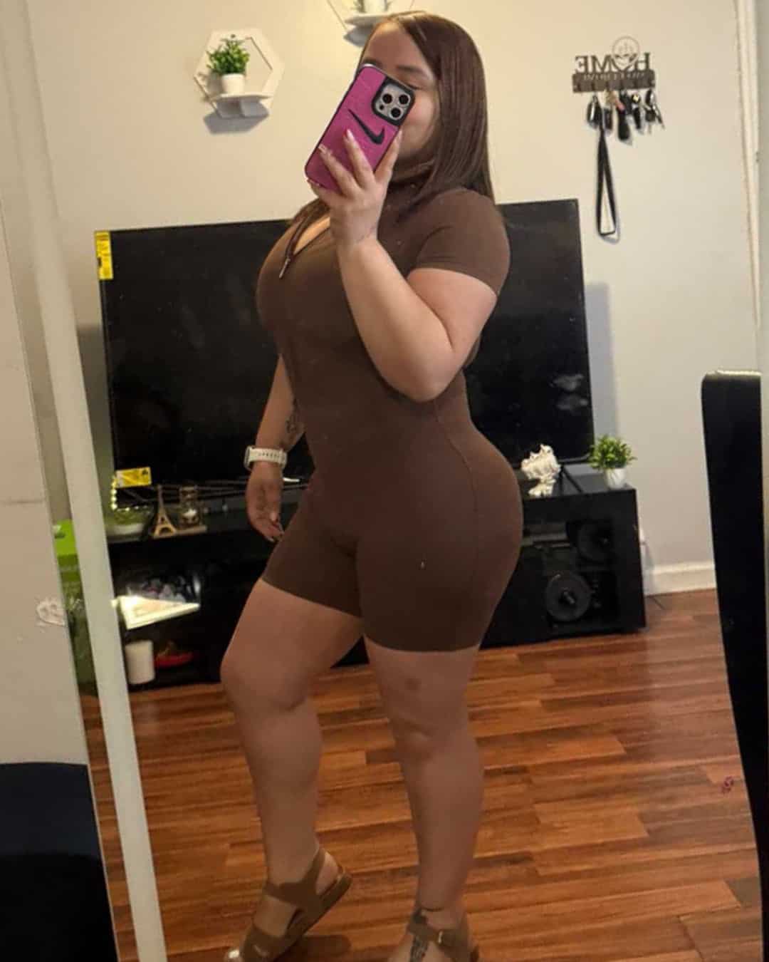 Curvy woman taking mirror selfie in brown athletic outfit at Nip Tuck Aesthetics clinic.